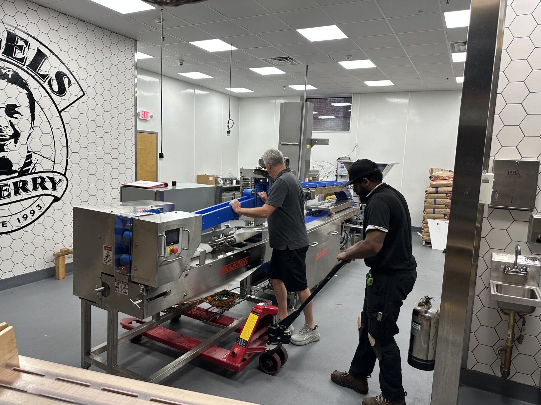 Two workers from BakTek Services operate and inspect a large stainless steel packaging machine in a clean, industrial kitchen setting with white tiled walls.
