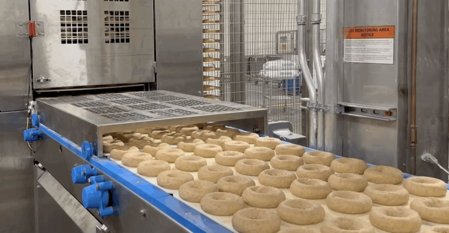 A conveyor belt carries rows of unbaked bagels into an industrial oven, while a Board Pusher & Rack Unloader helps streamline the baking process inside the commercial bakery facility.